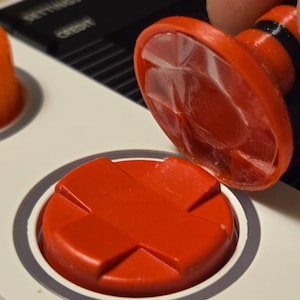 May include: Close-up of a red button with a cross-shaped design, next to a matching button being pressed. The buttons are on a white surface with a gray border. The button being pressed has a clear, textured top.