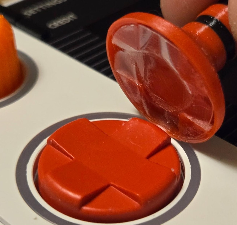 May include: Close-up of a red button with a cross-shaped design, next to a matching button being pressed. The buttons are on a white surface with a gray border. The button being pressed has a clear, textured top.