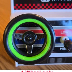 Puede incluir: Un volante negro con un borde verde. El volante tiene un diseño de radios plateados y un cubo central negro.