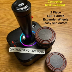 May include: Two black and red Atari GSP paddle expander wheels. The wheels are designed to easily slip on and off a joystick. The text "2 Piece GSP Paddle Expander Wheels easy slip on/off" is printed on a white background. The text "CONTROLLER NOT INCLUDED" is printed on a white sticker.