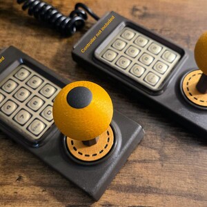 Puede incluir: Dos mandos de juego de estilo retro con un teclado numérico y un joystick. Los joysticks tienen una parte superior en forma de bola amarilla con un círculo negro. Los mandos son de color gris oscuro con el texto "Controller not included".
