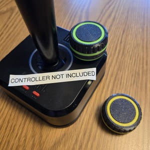 Może przedstawiać: Czarny joystick z zielono-czarną gałką i żółto-czarną gałką. Biała etykieta na joysticku głosi "KONTROLER NIE ZAWARTY W ZESTAWIE".