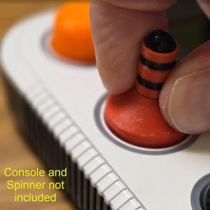 May include: Close-up of a red and black joystick button being pressed. The button is part of a white console with other orange buttons. The text "Console and Spinner not included" is visible in the image.