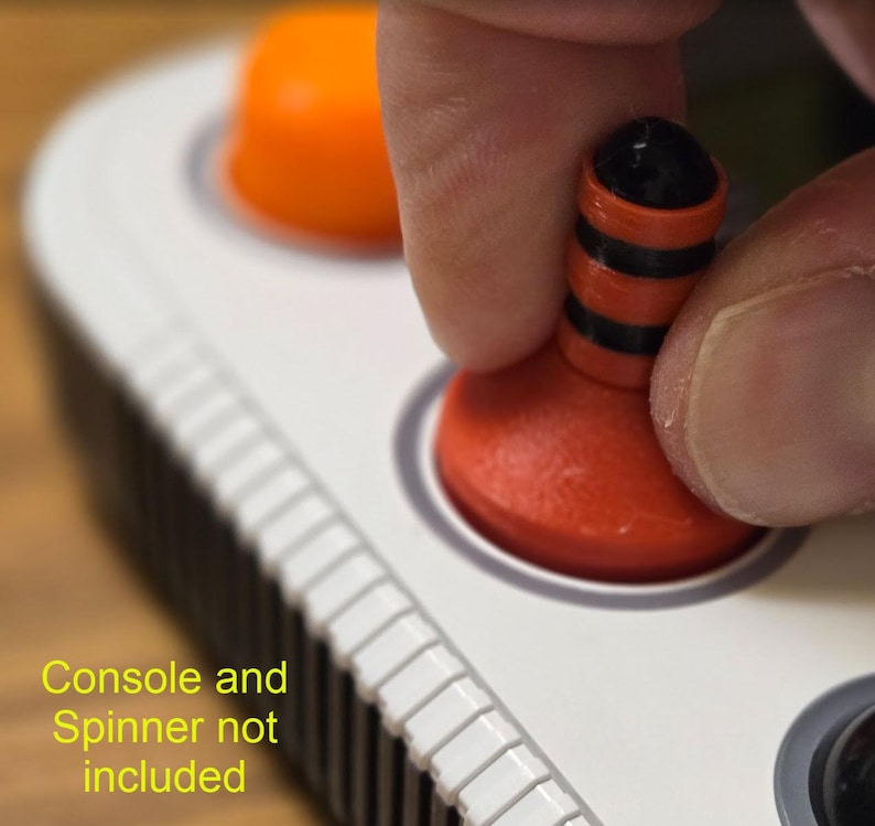 May include: Close-up of a red and black joystick button being pressed. The button is part of a white console with other orange buttons. The text "Console and Spinner not included" is visible in the image.