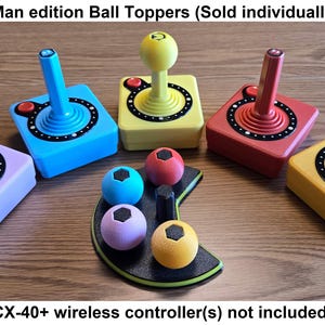 Pode incluir: Uma coleção de coloridos toppers de bolas CX-40+ edição Pac-Man. Os toppers estão em várias cores, incluindo azul, amarelo, vermelho e roxo. A imagem também mostra um suporte preto com toppers de bolas com tema Pac-Man. O texto na imagem diz "CX-40+ Pac-Man edition Ball Toppers".