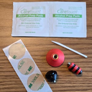 May include: An assortment of medical supplies and accessories, including two "CareTouch Alcohol Prep Pads" packets, adhesive circles, a red and gold object, a black object, a red and black striped object, and a toothpick.