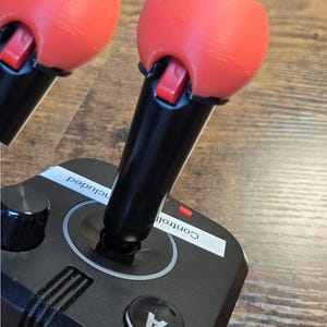 Könnte beinhalten: Ein schwarzer Joystick mit zwei roten Kugelaufsätzen. Der Joystick hat einen Knopf mit der Aufschrift "A" und einen Drehknopf. Der Text "Control" und "Included" ist auf der Basis des Joysticks sichtbar. Der Text "2 Ball set" ist in der oberen rechten Ecke des Bildes sichtbar.