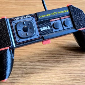 May include: Black Sega Genesis controller with red accents and a textured surface. The controller has a directional pad, two buttons, and a start button. The text "Controller NOT included" is printed on the controller.