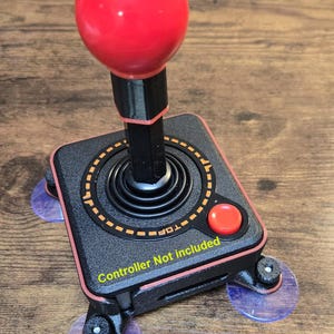 Könnte beinhalten: Schwarzer und roter Joystick-Controller mit einem roten, kugelförmigen Griff. Der Controller hat eine schwarze Basis mit einem orangefarbenen gepunkteten Kreis und einem roten Knopf. Der Text "Controller Not included" ist sichtbar.
