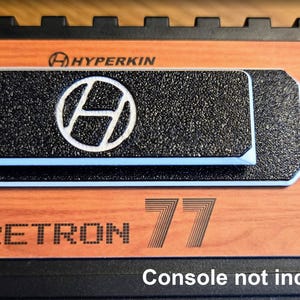 Peut inclure: Gros plan sur une console Retron 77. La console a une finition imitation bois avec un dessus noir texturé. Le logo Hyperkin est visible, ainsi que le texte "Retron 77". Le texte "Console non incluse" est également présent.