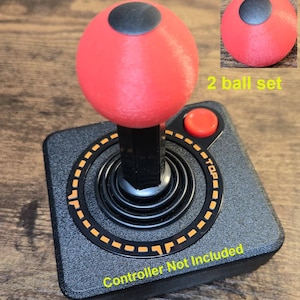 Puede incluir: Un mando de joystick negro y gris con una parte superior roja en forma de bola y un círculo negro en la parte superior. La imagen incluye una imagen más pequeña de una bola roja con un círculo negro. El texto dice "2 ball set" y "Controller Not Included".