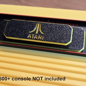 Puede incluir: Primer plano de una consola Atari 2600. La consola es amarilla con un panel de control negro. El logotipo de Atari es visible en una placa negra texturizada con ribete amarillo. El texto "2600+ console NOT included" está en la parte inferior.