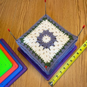 May include: A white and purple crochet square with a green border is being stretched on a purple plastic frame. The frame is sitting on a wooden surface with a yellow measuring tape.