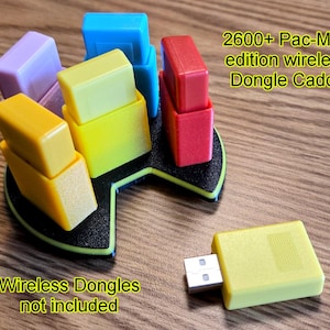 May include: A colorful Pac-Man edition wireless dongle caddy with multiple colored dongles. The caddy is black with a green trim. The text on the image reads "2600+ Pac-Man edition wireless Dongle Caddy" and "Wireless Dongles not included". A yellow dongle is shown.