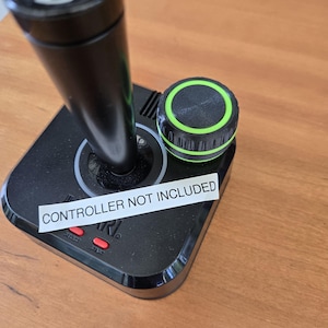 以下が含まれることがあります： 黒と緑の制御ノブが付いた黒いジョイスティックベース。ベースには「コントローラーは含まれていません」というステッカーが付いています。