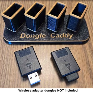 Könnte beinhalten: Ein Holzständer mit vier Schlitzen zum Aufbewahren kleiner elektronischer Geräte. Der Ständer ist schwarz mit Holzmaserung und trägt die Aufschrift "Dongle Caddy". Zwei USB-Funkadapter sind auf dem Bild zu sehen.