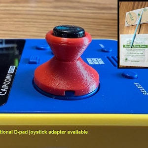 May include: A red joystick attachment installed on a blue and yellow handheld gaming console with the text CAPCOM EDITION. An inset image shows the red joystick component, three circular adhesive pads, a small tool, and an alcohol prep pad.