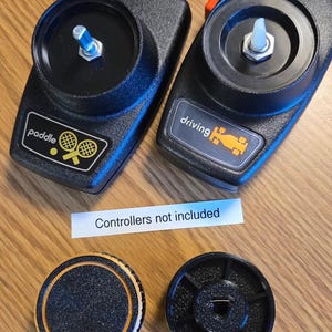 Könnte beinhalten: Zwei schwarze Atari-Gamecontroller mit orangenen Etiketten. Der linke Controller hat ein Etikett mit der Aufschrift "paddle" und einem Tennisschläger-Symbol. Der rechte Controller hat ein Etikett mit der Aufschrift "driving" und einem Autosymbol. Ein weißes Etikett unter den Controllern lautet "Controller nicht im Lieferumfang enthalten". Zwei schwarze runde Knöpfe mit einer strukturierten Oberfläche sind unter den Controllern zu sehen.