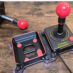 Puede incluir: Una consola de juegos Atari 2600 negra y roja con un joystick y un botón rojo. La consola tiene ventosas en la parte inferior. El texto "GET 2 ZONE" está impreso en el lateral de la consola. El texto "Controlador no incluido" está impreso en la consola y en un joystick separado.