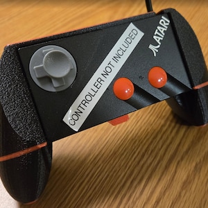 Puede incluir: Una consola de videojuegos Atari negra con un borde naranja. La consola tiene una almohadilla direccional gris, dos botones rojos y una etiqueta que dice "CONTROLADOR NO INCLUIDO".