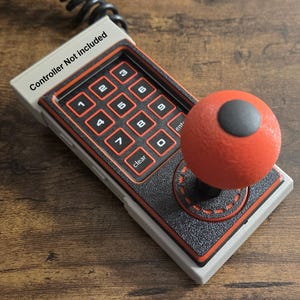 Puede incluir: Un mando de juego de estilo retro con un joystick rojo y un teclado numérico. El mando tiene una combinación de colores gris y negro con detalles en naranja. El texto "Controller Not included" es visible.