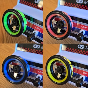 Puede incluir: Cuatro volantes de diferentes colores para un juego de carreras de coches de juguete. Los volantes son negros con un diseño de radios plateados y un anillo de color alrededor del exterior. Los colores son verde, rosa, azul y amarillo.