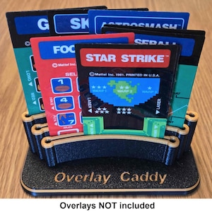 May include: A black plastic overlay caddy with the text "Overlay Caddy" printed on the front. The caddy is designed to hold multiple game overlays for vintage video game consoles. The overlays are not included.