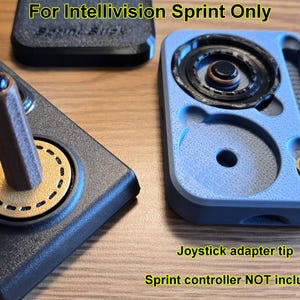 May include: An assortment of retro gaming accessories. A black controller with a brown joystick and gold accents is next to a blue component with circular parts. The text "For Intellivision Sprint Only" is visible.