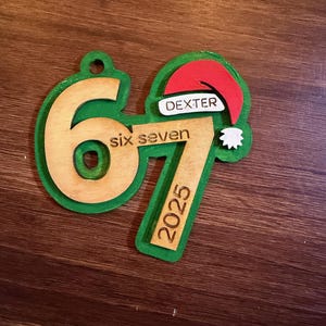 Puede incluir: Adorno navideño de madera con borde verde e interior de madera natural. El adorno presenta el número "67" y las palabras "six seven" y "2025". También se incluye un pequeño gorro de Papá Noel con el nombre "DEXTER".
