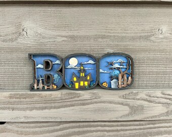 May include: A wooden Halloween decoration spelling "BOO". Each letter features a spooky scene: a ghost, haunted house, and graveyard. The letters are outlined in black with a blue background and a wooden fence.