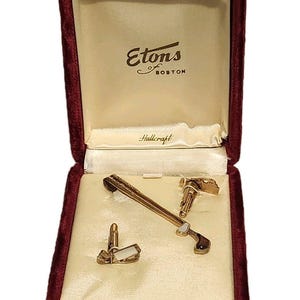 May include: A gold-toned tie bar and two matching cufflinks with white rectangular inlays, presented in a deep red velvet-lined box. The interior lid features the text Etons of Boston and Hallcraft printed in black on a cream satin fabric.