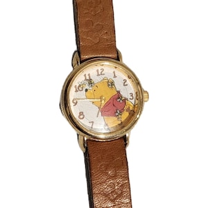 May include: A gold-toned wristwatch with a brown leather band. The watch face features a cartoon image of Winnie the Pooh with bees. The watch has a round face with black numerals and hands.