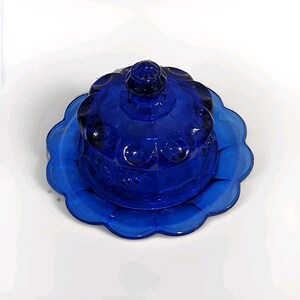 May include: A cobalt blue glass butter dish featuring a domed lid with a circular knob and a scalloped edge base. The lid is decorated with a repeating pattern of raised circular indentations, creating a textured surface across the glass.