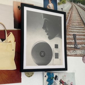 Może przedstawiać: Czarno-biały wydruk w ramce przedstawiający profil kobiety z butelką perfum Chanel i okrągłym pudełkiem. Pudełko ma logo Chanel.