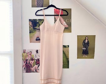 May include: A pale pink slip dress with a lace trim at the hem. The dress is hanging on a black hanger against a white wall.