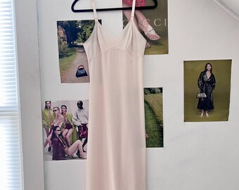 May include: A pale pink slip dress with thin straps and a V-neckline. The dress is hanging on a black hanger against a white wall.