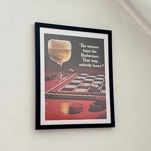 Może przedstawiać: Wizerunek vintage'owego ogłoszenia piwa Budweiser z szklanką piwa i szachownicą z tekstem "The winner buys the Budweiser. That way, nobody loses*"