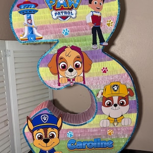 May include: A colorful number three piñata featuring characters from Paw Patrol. The piñata has a blue glitter border and includes the Paw Patrol logo, cartoon images of Chase, Skye, Marshall, and Ryder, and the name "Caroline".