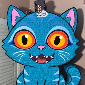May include: A large, blue cat-shaped piñata with a textured surface. The cat has large, orange and yellow eyes, pink ears, and a smiling mouth. A small, black decorative piece sits on top of the cat's head.
