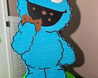 May include: A large, blue Cookie Monster piñata with a cookie in its mouth. The piñata has large white eyes with black pupils and a black mouth. The base is green, and a sign reads "123 BRYCE".