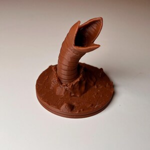 May include: A brown, 3D-printed sculpture of a sandworm emerging from a circular base. The worm's open mouth and segmented body are detailed. The base has a textured, rocky appearance. The sculpture is set against a white background.