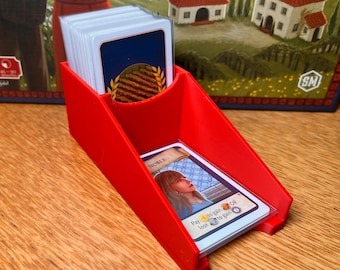 May include: A red plastic card holder with a slanted design, holding a stack of game cards. One card is visible, featuring a portrait of a woman and the word "NOBLE". The cards have a blue border and gold accents. The background shows a board game.