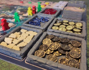 May include: A board game setup with various components. Includes wooden player pieces in red, yellow, and green, along with trays of tokens in different colors and shapes, and a deck of cards. The game board is visible in the background.