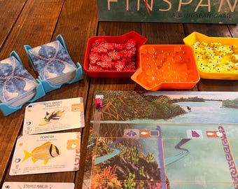 May include: A board game setup with a game board, cards, and various game pieces in red, orange, and yellow trays. The game box reads "FINSPAN A WINGSPAN GAME". Blue card holders hold stacks of cards with a blue and white design.