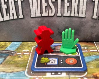 May include: A board game scene with a red wooden figure and a green hand-shaped piece on a blue game card. The card features a house icon, a tree, and the number 2. The game board has a river and road design.