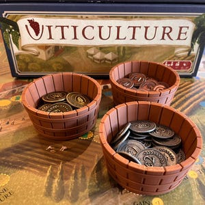 Puede incluir: Tres recipientes de madera marrón con forma de barril llenos de fichas de juego están sobre un juego de mesa. Las fichas son de varios colores, incluyendo oro y plata. La caja del juego, etiquetada "Viticuture", está en el fondo.