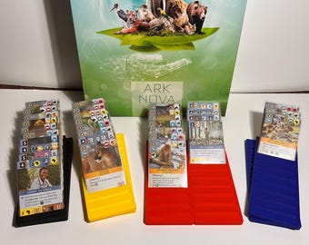 May include: A board game box with an elephant and other animals on a green background. Several stacks of cards are in front of the box, each in a different colored holder: black, yellow, red, and blue. The game is called Ark Nova.