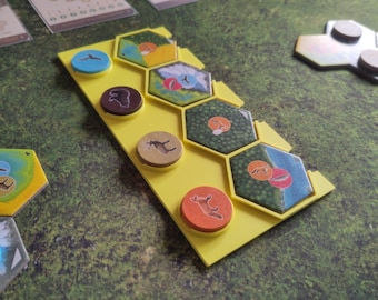 May include: A yellow game board with hexagonal tiles and circular tokens. The tiles feature illustrations of animals and landscapes. The tokens are blue, brown, tan, and orange, each with a different animal illustration. The game pieces are on a green surface.
