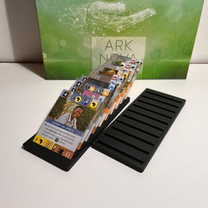 K&ouml;nnte beinhalten: Ein Brettspiel-Setup mit einem Stapel illustrierter Karten in einem schwarzen Halter, mit der Spielbox im Hintergrund. Auf der Box steht "ARK NOVA". Die Karten haben Symbole und Text, darunter "EXPERT ON AFRICA".