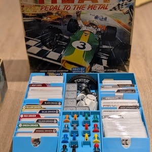 Könnte beinhalten: Brettspiel "HEAT: Pedal to the Metal" mit einer Box, die eine Rennwagen-Illustration zeigt. Das Spiel enthält Karten, Fahrer-Token und Spielsteine, die alle in einem blauen Behälter aufbewahrt werden. Der Text "DAYS OF WONDER" ist sichtbar.